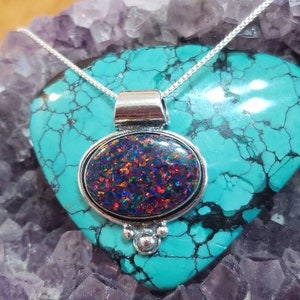 May include: A silver pendant necklace with a round, iridescent opal gemstone set in a silver bezel. The pendant is on a turquoise background.