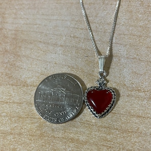 May include: A silver chain necklace with a red carnelian heart pendant. The pendant is set in a silver frame with a detailed design. The necklace is about the size of a US quarter.