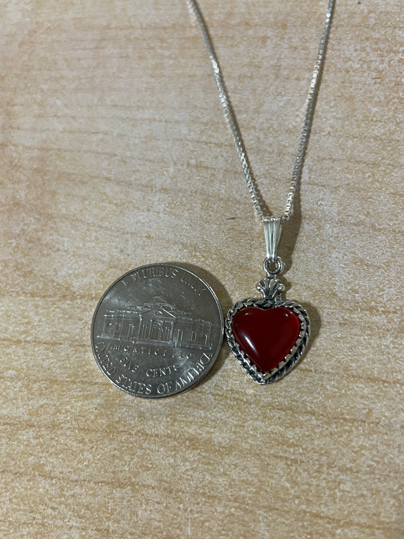 May include: A silver chain necklace with a red carnelian heart pendant. The pendant is set in a silver frame with a detailed design. The necklace is about the size of a US quarter.