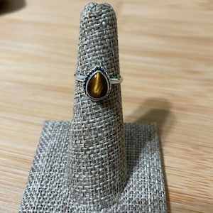 Puede incluir: Un anillo de plata con una piedra de ojo de tigre en forma de lágrima. El anillo tiene un borde de cuentas alrededor de la piedra.
