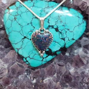 May include: A silver chain necklace with a heart-shaped pendant featuring a dark iridescent opal stone. The necklace is displayed on a turquoise and purple crystal background.