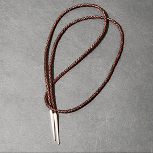 May include: A brown braided leather bolo tie with silver-colored tips. The bolo tie is displayed on a dark gray surface. The cord is looped and knotted, with the tips hanging down.