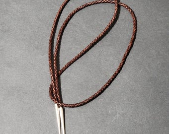 May include: A brown braided leather bolo tie with silver-colored tips. The bolo tie is displayed on a dark gray surface. The cord is looped and knotted, with the tips hanging down.
