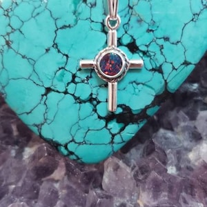 May include: A silver cross pendant with a round, iridescent opal gemstone in the center. The pendant is on a silver chain and is resting on a turquoise stone.