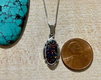 May include: A silver chain necklace with a pendant featuring a black oval opal with iridescent rainbow colors. The pendant is set in a silver frame with a decorative design. The necklace is shown next to a US penny for size comparison.