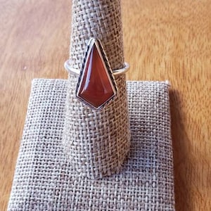 May include: A silver ring with a red carnelian gemstone set in a geometric shape. The ring is displayed on a fabric ring holder.