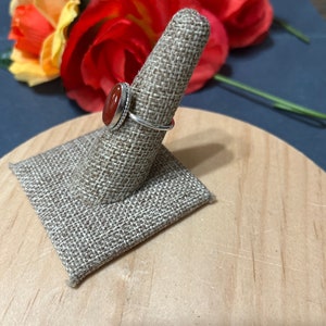 May include: A silver ring with a red carnelian stone set in a bezel setting. The ring is displayed on a fabric ring holder.