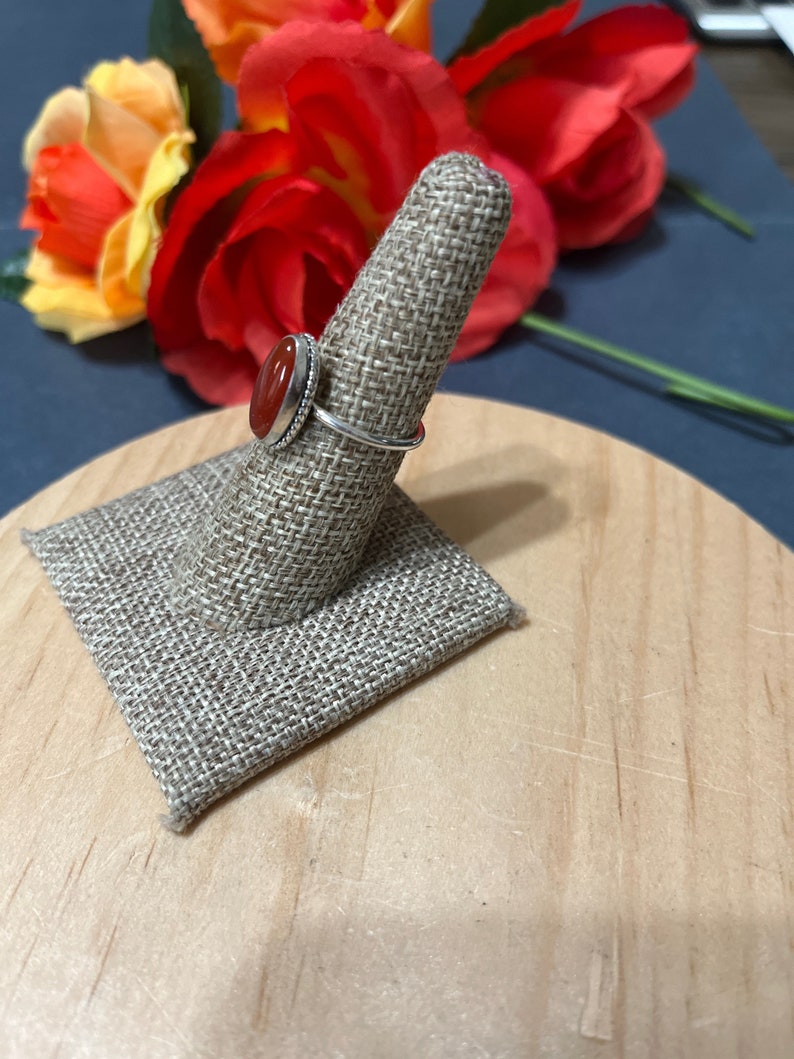 May include: A silver ring with a red carnelian stone set in a bezel setting. The ring is displayed on a fabric ring holder.