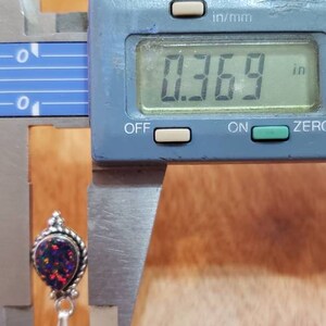 May include: A close-up of a silver pendant with a teardrop-shaped opal gemstone. The opal is iridescent with shades of blue, green, and red. The pendant is measured with a digital caliper, showing a measurement of 0.369 inches.