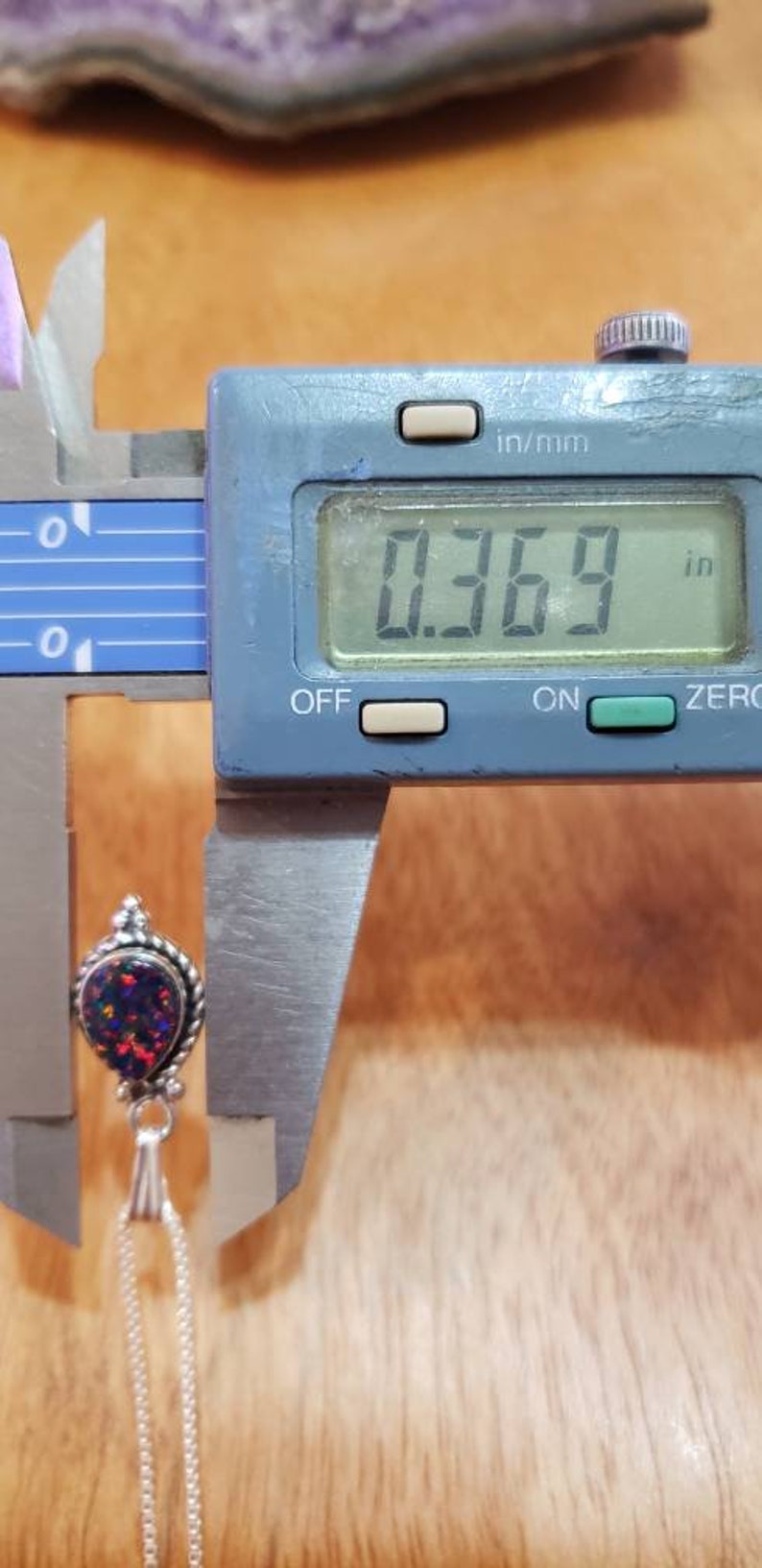 May include: A close-up of a silver pendant with a teardrop-shaped opal gemstone. The opal is iridescent with shades of blue, green, and red. The pendant is measured with a digital caliper, showing a measurement of 0.369 inches.