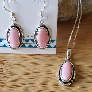 May include: A silver jewellery set comprising a necklace and earrings. Each piece features an oval, pale pink stone set in a decorative silver frame. The earrings are drop style, and the necklace has a fine chain.