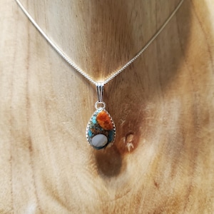 May include: A teardrop-shaped pendant necklace featuring a mosaic inlay of orange, turquoise, and white stone fragments set in a silver-toned metal frame. The pendant hangs from a thin, silver-toned chain, showcasing a polished, multi-colored surface.