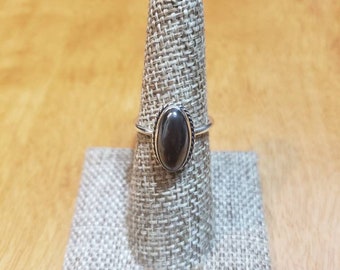 Puede incluir: Un anillo de plata con una piedra ovalada negra engastada en un engaste de bisel. El anillo está en un expositor de anillo de tela.