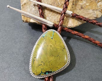 May include: A bolo tie with a green and blue turquoise stone set in silver. The bolo tie is on a brown leather cord.