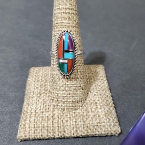 Op de afbeelding: Een zilveren ring met een ovale steen die in het midden is gezet. De steen is gemaakt van meerdere kleuren turquoise, koraal en andere stenen. De ring ligt op een bruine stoffen ringstandaard.