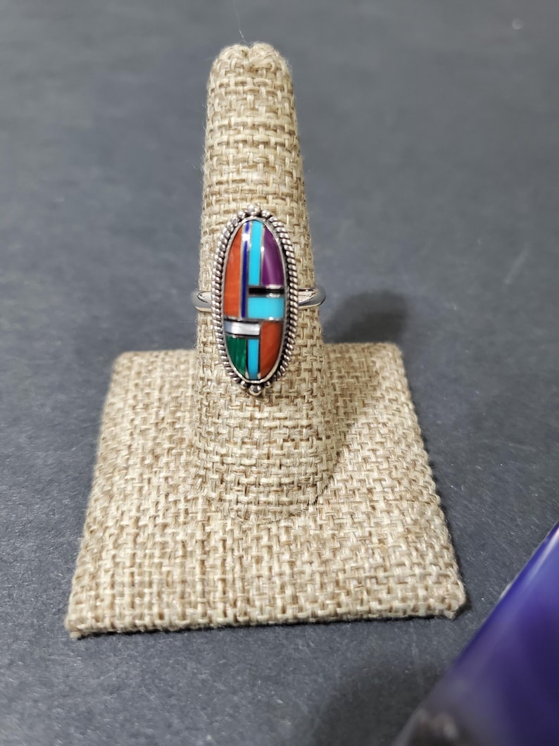 Op de afbeelding: Een zilveren ring met een ovale steen die in het midden is gezet. De steen is gemaakt van meerdere kleuren turquoise, koraal en andere stenen. De ring ligt op een bruine stoffen ringstandaard.