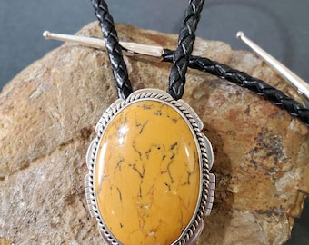 May include: A bolo tie with a large, oval-shaped, yellow gemstone set in a silver frame. The bolo tie is made with a black leather cord.