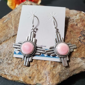 May include: A pair of silver earrings with pink stone inlays, shaped like a stylized sun symbol. The earrings are on a white card with the word "Silver" printed on it.