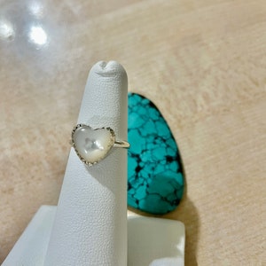 May include: A silver ring with a white heart-shaped stone. The ring is on a white ring display stand.