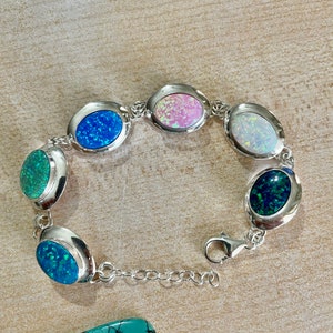 May include: A silver bracelet featuring oval-shaped opal stones in various colors, including blue, pink, green, and white. The bracelet has a lobster clasp closure and is displayed on a light brown surface.