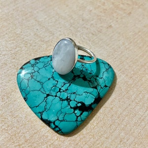 Puede incluir: Un anillo de plata con una piedra ovalada blanca. El anillo est&aacute; apoyado en una piedra turquesa con vetas negras.