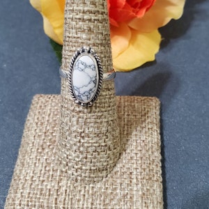May include: A silver ring with a white oval-shaped stone set in a silver bezel. The stone has a marbled pattern. The ring is displayed on a brown fabric ring holder.