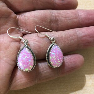 May include: A pair of silver dangle earrings with pink opal teardrop stones. The stones are set in a silver bezel with a rope-like border.