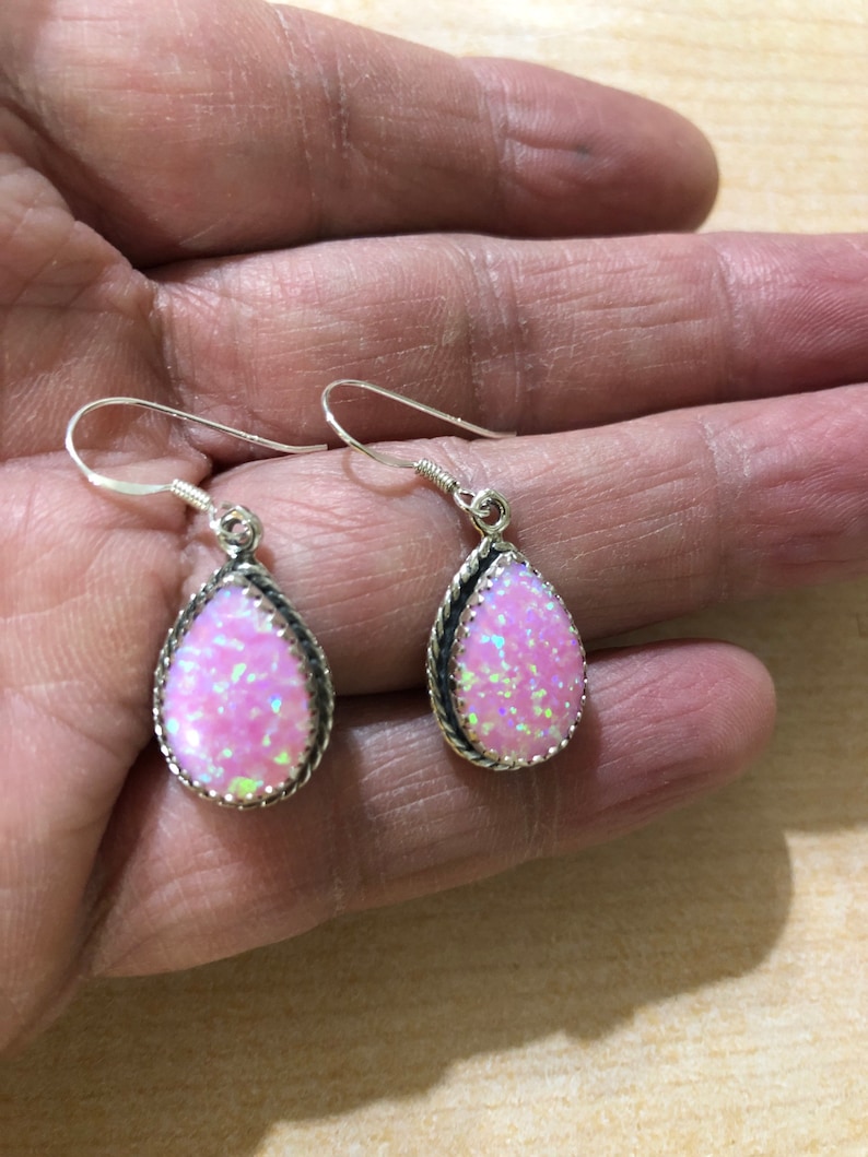 May include: A pair of silver dangle earrings with pink opal teardrop stones. The stones are set in a silver bezel with a rope-like border.