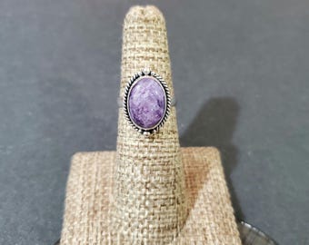 Op de afbeelding: Een zilveren ring met een paarse ovale edelsteen gezet in een zilveren bezel. De ring staat op een bruine stoffen ringstandaard.