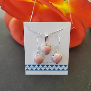 May include: A jewellery set featuring a sterling silver chain necklace and two matching drop earrings, each with a smooth, light pink heart-shaped stone pendant. The items are displayed on a white card with the text Sterling Silver printed on it.