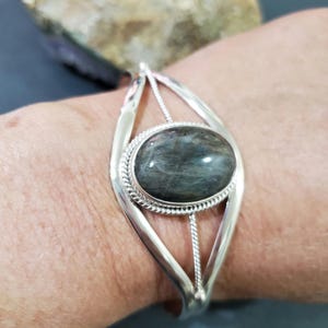 May include: Silver-toned cuff bracelet featuring an oval, iridescent gemstone. The gemstone is set in a detailed bezel, and the bracelet has an open design with decorative elements. The bracelet is worn on a wrist.