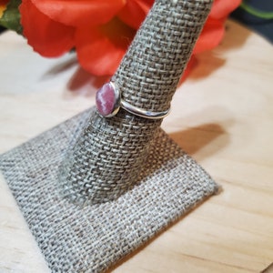 Puede incluir: Un anillo de plata con una piedra preciosa rosa y blanca engastada en un engaste de bisel. El anillo se muestra en un soporte de anillo de tela.