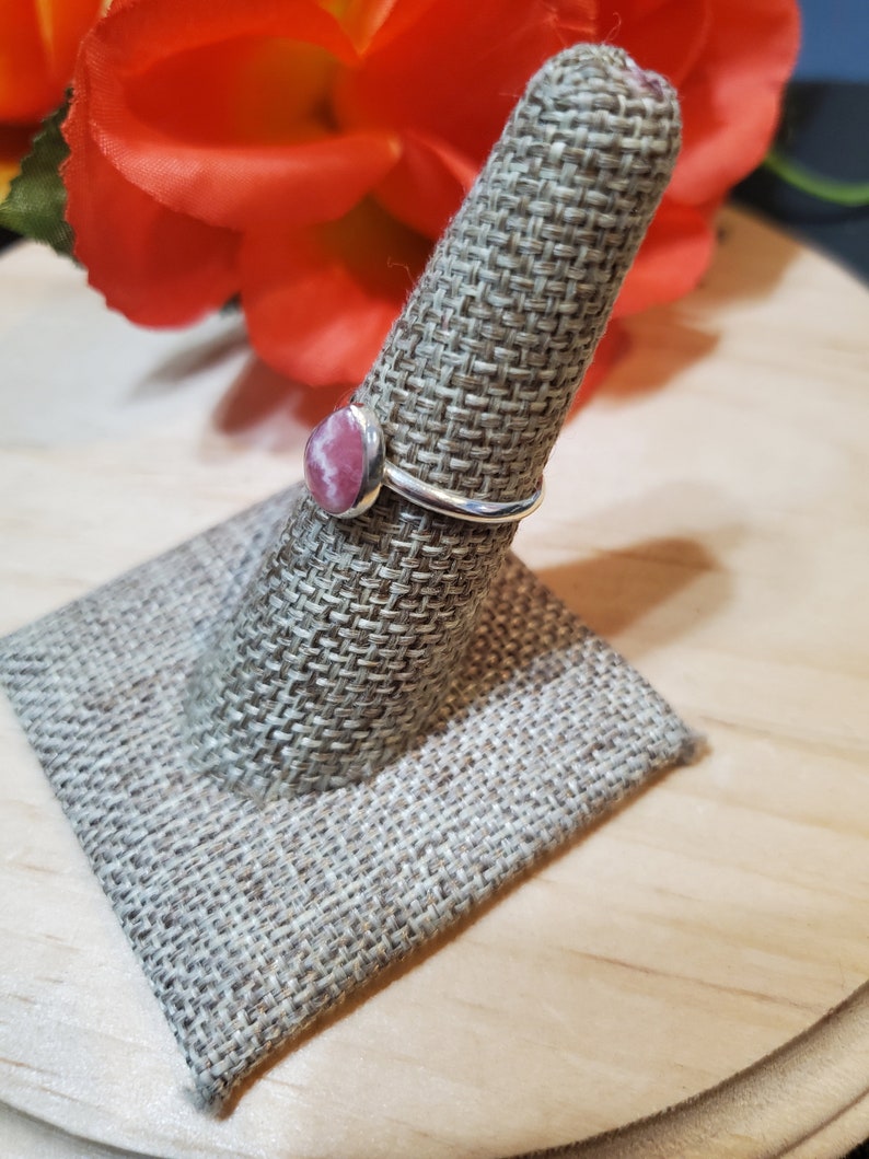 Puede incluir: Un anillo de plata con una piedra preciosa rosa y blanca engastada en un engaste de bisel. El anillo se muestra en un soporte de anillo de tela.