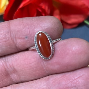 May include: A silver ring with an oval-shaped carnelian gemstone. The ring has a delicate, detailed design around the stone.