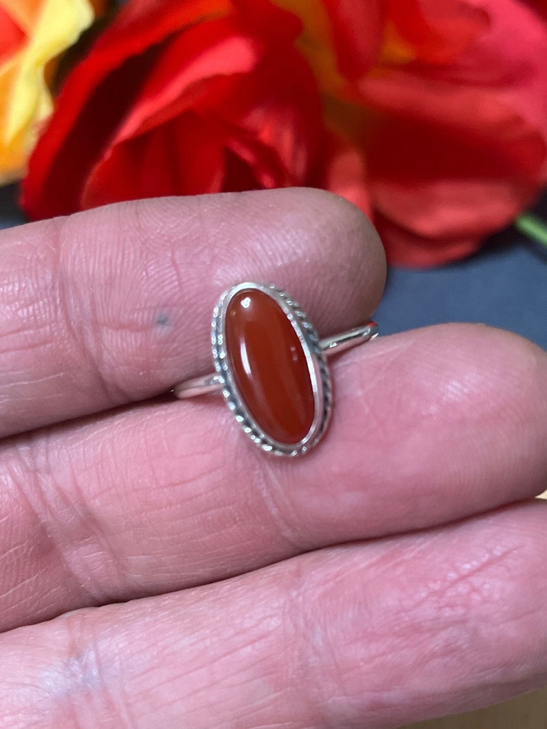 May include: A silver ring with an oval-shaped carnelian gemstone. The ring has a delicate, detailed design around the stone.