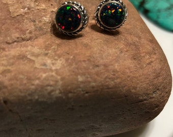 May include: A pair of round stud earrings with black opal stones. The opals have flecks of green, orange, and red. The earrings are set in a silver-colored metal with a twisted rope design around the edge. The earrings are displayed on a brown stone.