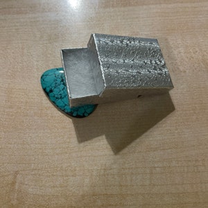 May include: A silver gift box with a textured pattern, open to reveal a turquoise-colored stone inside.