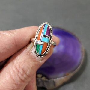 Op de afbeelding: Een zilveren ring met een ovale steen in het midden. De steen is gemaakt van turquoise, koraal en andere gekleurde stenen.