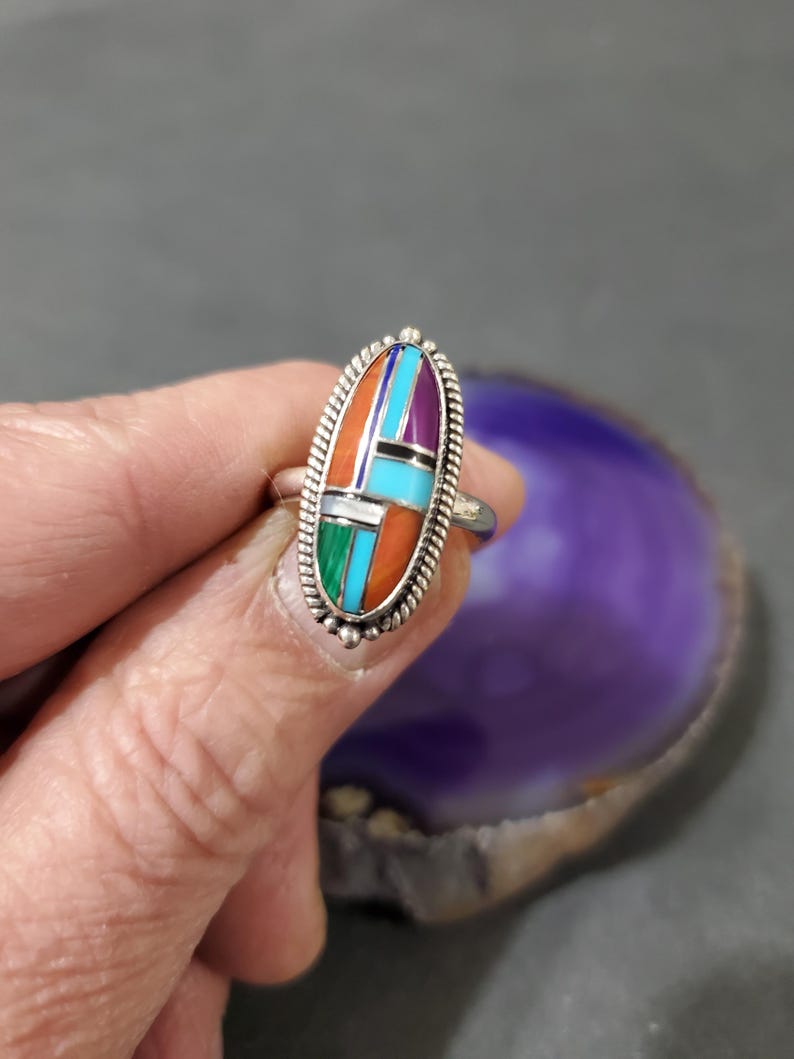 Op de afbeelding: Een zilveren ring met een ovale steen in het midden. De steen is gemaakt van turquoise, koraal en andere gekleurde stenen.