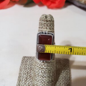 May include: A silver ring with a rectangular red carnelian stone. The ring is on a fabric ring holder and a measuring tape is next to it.