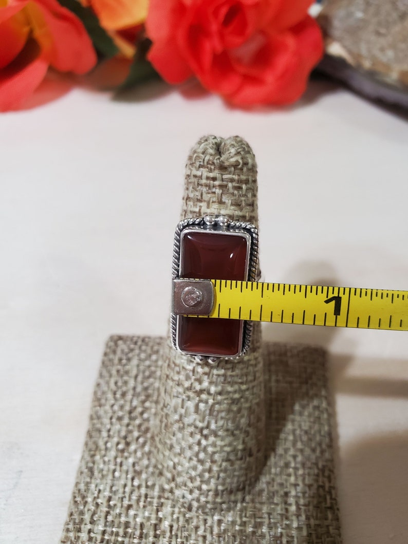 May include: A silver ring with a rectangular red carnelian stone. The ring is on a fabric ring holder and a measuring tape is next to it.