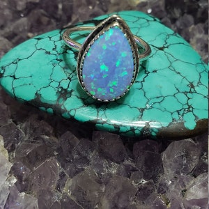 May include: A silver ring with a teardrop-shaped opal gemstone. The opal is a light blue color with green and blue flashes. The ring is set on a bed of purple amethyst crystals.