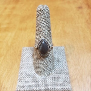 May include: A silver ring with a dark gray teardrop-shaped stone set in a silver bezel. The ring is on a fabric ring display stand.