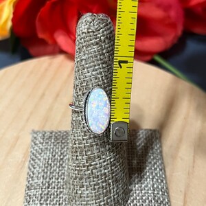 May include: A silver ring with an oval-shaped opal gemstone. The opal is iridescent with shades of white, pink, and blue. The ring is displayed on a fabric ring holder.