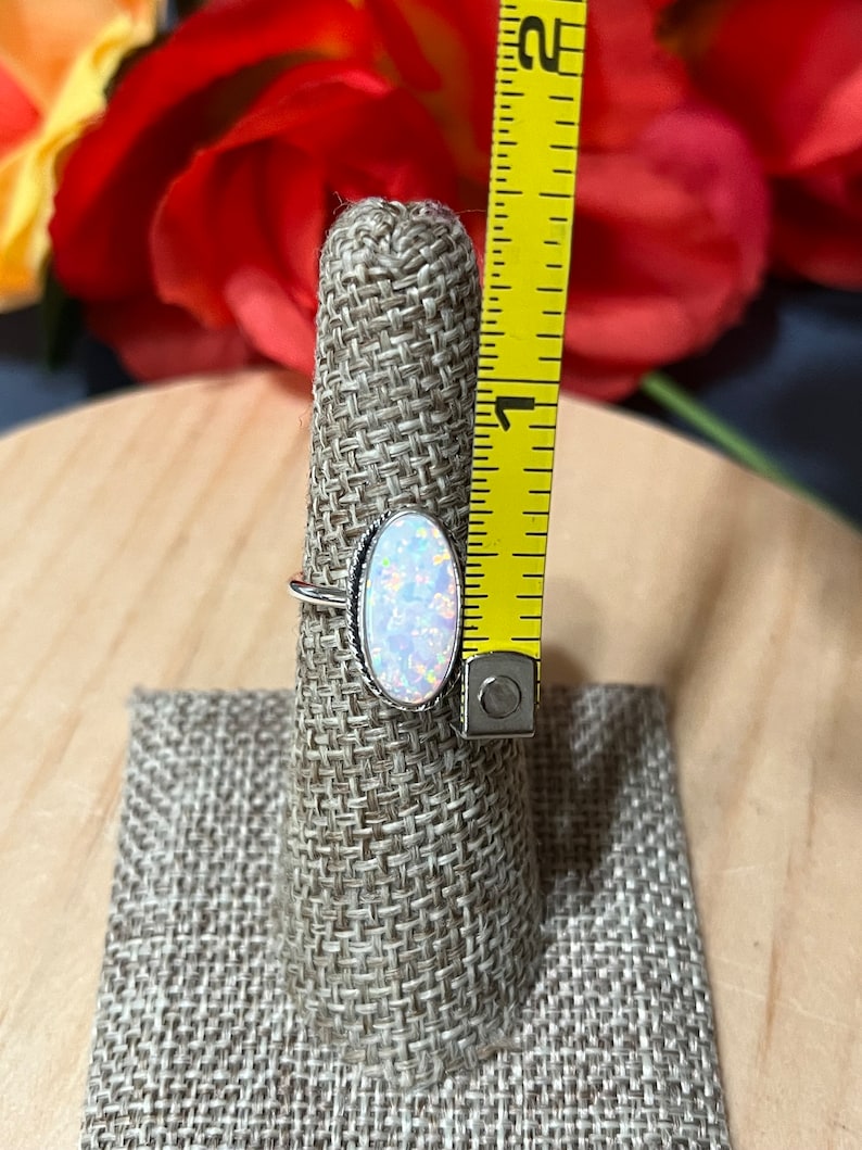 May include: A silver ring with an oval-shaped opal gemstone. The opal is iridescent with shades of white, pink, and blue. The ring is displayed on a fabric ring holder.