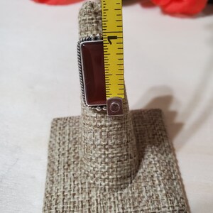 May include: A silver ring with a rectangular, dark red gemstone. The ring is displayed on a brown fabric ring holder.