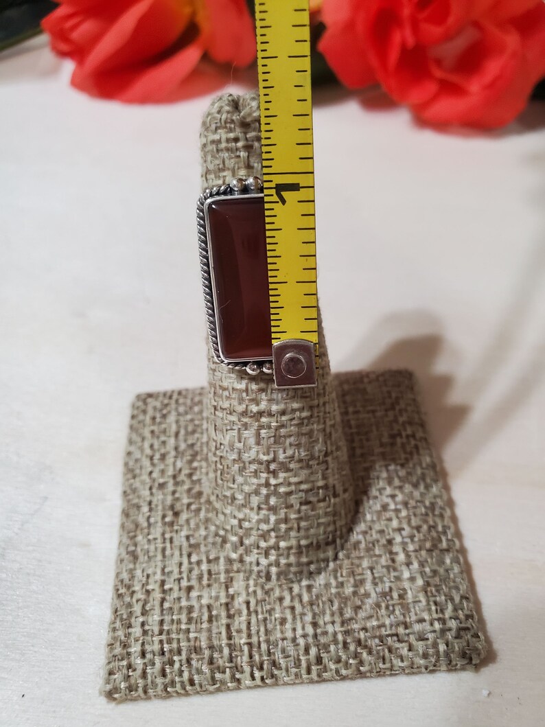 May include: A silver ring with a rectangular, dark red gemstone. The ring is displayed on a brown fabric ring holder.