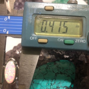 May include: A close-up of a white opal gemstone set in silver, measured at 0.415 inches, with a turquoise cabochon in the background.