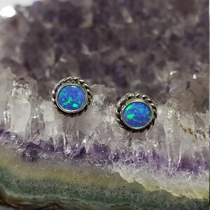 Puede incluir: Un par de pendientes de plata con piedras de ópalo azul engastadas en un diseño de cuerda. Los pendientes se muestran en una geoda de amatista púrpura.