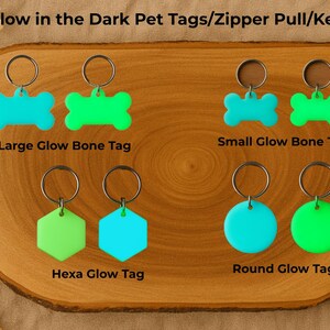 Pu&ograve; includere: Un espositore in legno presenta vari tag per animali domestici e portachiavi che si illuminano al buio. I tag sono a forma di osso, esagono e rotondi, in blu e verde. Il testo recita "Glow in the Dark Pet Tags/Zipper Pull/Key Fobs" e "It Glows".